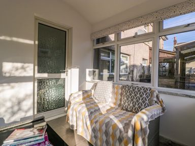 A sunlit sitting area with a sofa covered by a patterned throw and cushions next to windows and a textured glass door at Rose Cottage in Bream