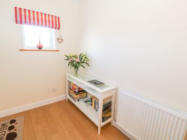 A corner of a room with a small white bookshelf holding board games and books with a vase of flowers on top next to a window and a radiator at Llain Bach in Llangefni