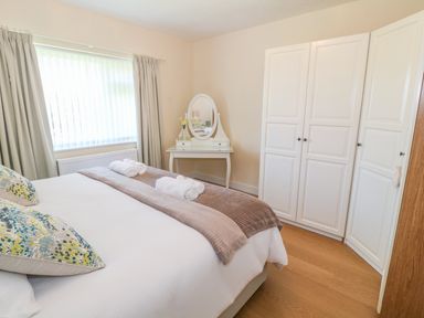 A bedroom with a large bed with pillows and folded towels next to a window with blinds and curtains a dressing table with mirror and wardrobe with three white doors at Llain Bach in Llangefni