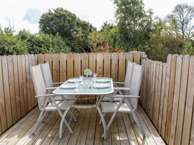 An outdoor dining area with a glass table set with plates and glasses surrounded by six beige chairs on a wooden deck with wooden fencing and trees in the background at Llain Bach Llangefni