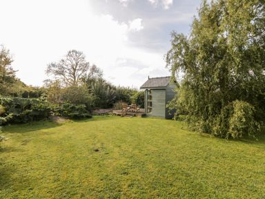 A garden with a small shed and two chairs on a wooden deck surrounded by trees and bushes at Llain Bach in Llangefni