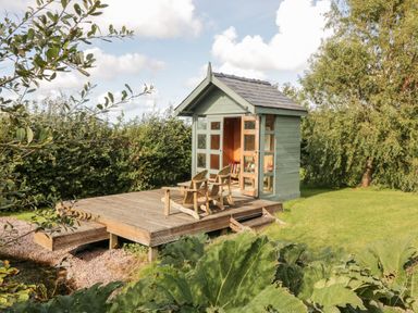 A small green garden shed with open doors on a wooden deck with two wooden chairs surrounded by trees and grass at Llain Bach in Llangefni