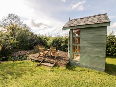 A small green wooden shed with glass windows beside a wooden deck with two chairs and a table in a garden at Llain Bach in Llangefni