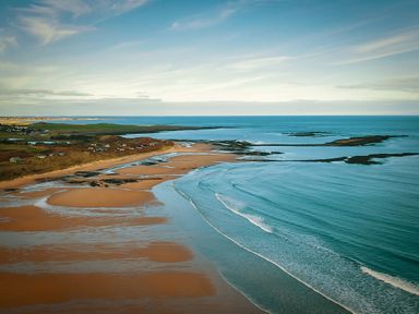 A coastal beach with sand, scattered rocks, and small waves near a shoreline with houses and green fields at Oystercatcher in Embleton