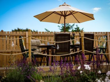 An outdoor seating area with a table and chairs under a large umbrella surrounded by a wooden fence and purple flowers at Oystercatcher in Embleton