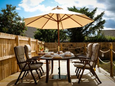 An outdoor wooden dining table with six cushioned chairs and a large umbrella in a fenced yard