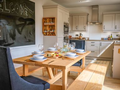 A dining area with a wooden table set with bowls, plates, wine glasses and a cheese board next to a kitchen with cream cabinets and appliances at Oystercatcher in Embleton