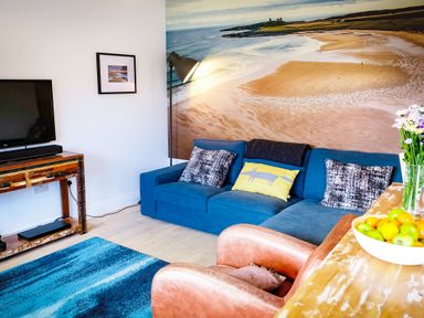 A living room with a blue sofa a wooden TV stand a brown chair and a bowl of fruit at Oystercatcher in Embleton