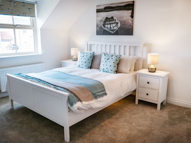 A bedroom with a white bed frame blue throw pillows two white bedside tables and a window at Oystercatcher in Embleton