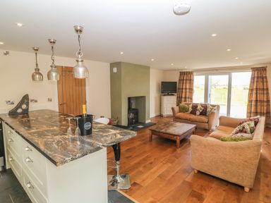 An open living room and kitchen area with a marble countertop island three pendant lights two beige sofas a wooden coffee table a fireplace and large sliding glass doors at Rhos Y Foel Cottage in Nefyn