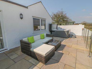 An outdoor patio area with a wicker sofa, green cushions, a glass coffee table, and a hot tub at Rhos Y Foel Cottage in Nefyn