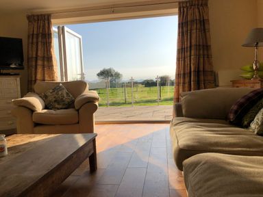 A living room with beige armchairs, a wooden coffee table, curtains, and an open glass door showing a green field outside at Rhos Y Foel Cottage in Nefyn
