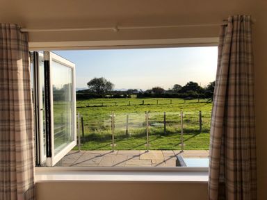 A window with plaid curtains open to a green field and glass railing at Rhos Y Foel Cottage in Nefyn