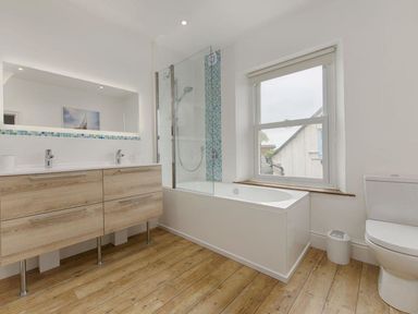 A bathroom with a wooden cabinet with two sinks a bathtub with a glass shower panel a window a toilet and wooden floor at Anchorage in Watchet
