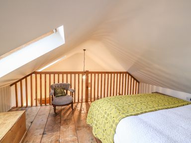 An attic bedroom with a bed covered by a yellow patterned blanket a wooden chest a gray armchair with a cushion and a wooden railing at Bwthyn Lan in Boncath