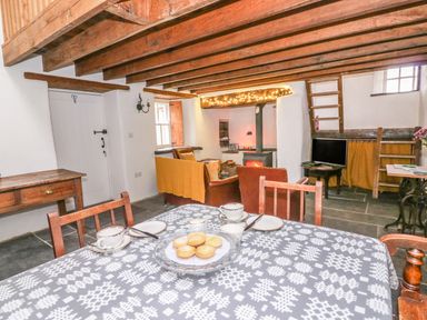 A dining area with a table set for tea and a living room with a wood burner stove and seating under wooden ceiling beams at Bwthyn Lan in Boncath