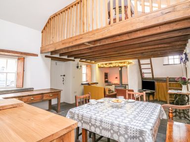 A dining area with a table and chairs under a wooden loft with a sitting area and wood burning stove at Bwthyn Lan in Boncath