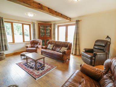A living room with leather sofas a wooden coffee table a rug a wooden cabinet and windows with curtains at Pentrego Isaf in Meifod
