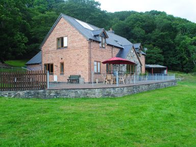 A brick and stone house with a patio and outdoor furniture including a red umbrella surrounded by grass and trees at Pentrego Isaf in Meifod