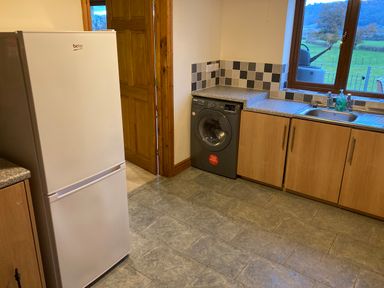 A kitchen with a white fridge freezer washing machine wooden cabinets and a sink with a window overlooking a field at Pentrego Isaf in Meifod