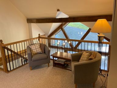 A seating area with two armchairs a wooden table and a floor lamp on an upper level with railing and large window at Pentrego Isaf in Meifod