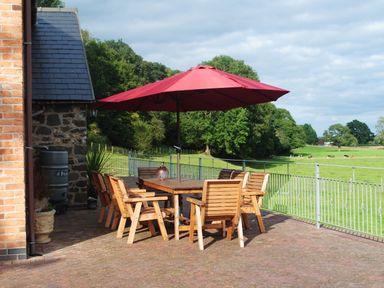Outdoor patio with wooden table and chairs under a red umbrella overlooking a green field at Pentrego Isaf in Meifod