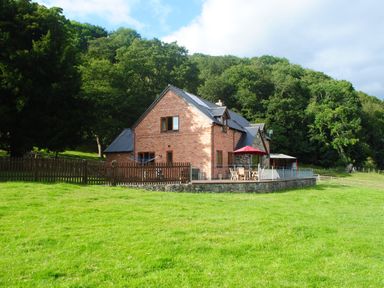 A brick house with a patio and red umbrella in a grassy field near trees at Pentrego Isaf in Meifod