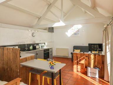 A kitchen and dining area with a table and yellow chairs a wooden partition a sofa and a wooden counter with a monitor at The Daffodil Shed in Bere Alston