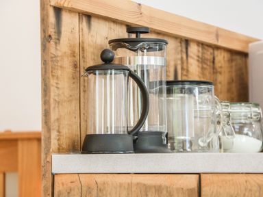 Two glass French presses a glass mug and glass jars with white contents on a kitchen counter with wooden paneling at The Daffodil Shed in Bere Alston