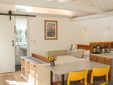 A bedroom with a bed a small table with two yellow chairs a cabinet with baskets and a small bathroom visible through a sliding door at The Daffodil Shed in Bere Alston
