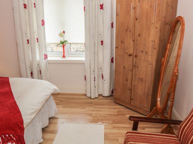 A bedroom with a bed partly covered by a red throw a wooden wardrobe a standing mirror a striped armchair and curtains with flower patterns at Cnoc suan in Aughavas County Leitrim