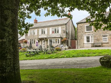 A stone inn with outdoor picnic tables and umbrellas next to a grassy area framed by tree branches at The Smithy in Blisland