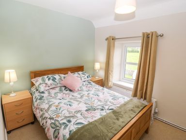 A bedroom with a double bed and floral bedding with two bedside tables and lamps near a window with curtains at 3 Green Terrace in Llangaffo near Newborough