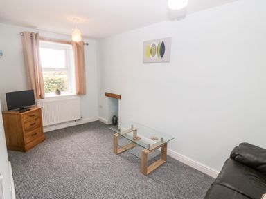 A small living room with a glass coffee table a black leather sofa a wooden TV stand with a small TV and a window with curtains at 3 Green Terrace in Llangaffo near Newborough