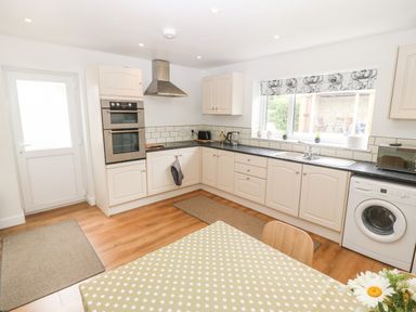 A kitchen with white cabinets a washing machine oven and a dining table with a polka dot tablecloth at 3 Green Terrace in Llangaffo near Newborough