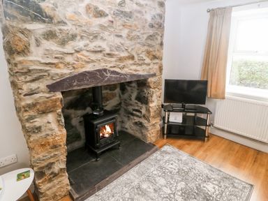 A room with a stone fireplace and wood burning stove and a television on a stand next to a window at 3 Green Terrace in Llangaffo near Newborough