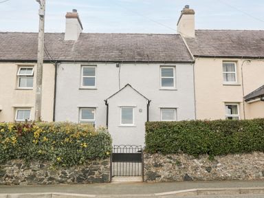A stone cottage with white walls and two chimneys behind a stone wall and garden hedge at 3 Green Terrace in Llangaffo near Newborough