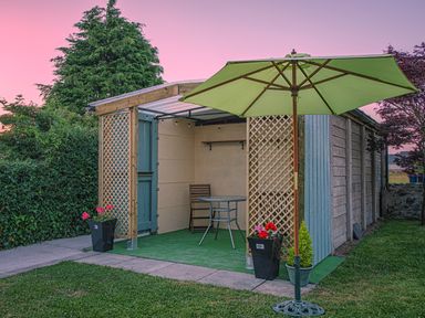 A garden shed with a green patio umbrella a table one chair and flower pots at 3 Green Terrace in Llangaffo near Newborough