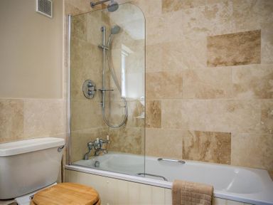 A bathroom with a bathtub shower combo a glass shower screen a toilet with a wooden seat and beige tiled walls at Lime Tree in Burford
