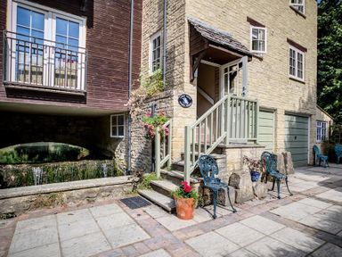 An outdoor patio area with stone steps a railing several chairs potted plants and a building beside a water feature at Lime Tree in Burford