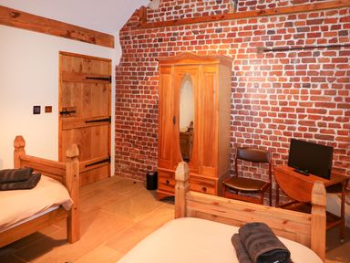 A bedroom with two wooden beds a wooden wardrobe a chair and a small table with a television against a brick wall at Honey Buzzard Barn in Fakenham