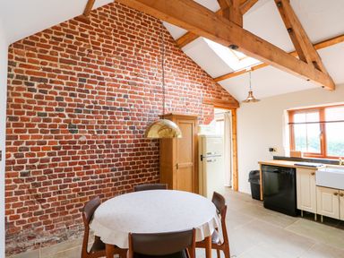 A kitchen with a round table and chairs near a brick wall and wooden ceiling beams at Honey Buzzard Barn in Fakenham