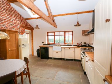 A kitchen with wooden beams cream cabinets a round dining table and brick wall at Honey Buzzard Barn in Fakenham