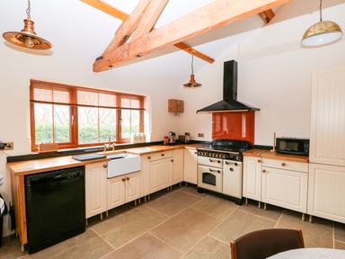 A kitchen with wooden countertops white cabinets a black dishwasher a farmhouse sink a stove with a red backsplash and wooden ceiling beams at Honey Buzzard Barn in Fakenham