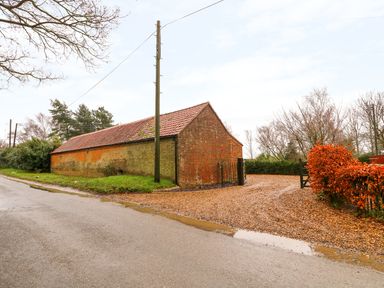 A brick barn with a tiled roof next to a gravel driveway and trees at Honey Buzzard Barn in Fakenham