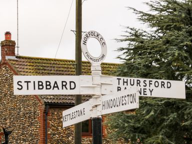 A white metal signpost with directions to Stibbard Thursford Barney Kettlestone and Hindolveston in a village setting with a stone house and tree in the background at Honey Buzzard Barn in Fakenham