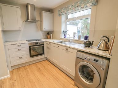 A kitchen with white cabinets a stainless steel oven washing machine and a window with a patterned blind at Ty Taid in Rhosneigr