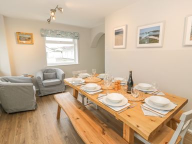 A dining room with a wooden table set for six, two grey armchairs, and framed pictures on the wall at Ty Taid in Rhosneigr