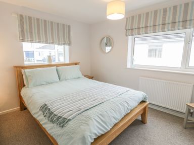 A bedroom with a wooden bed with light blue bedding two windows with striped blinds and a round mirror on the wall at Ty Taid in Rhosneigr