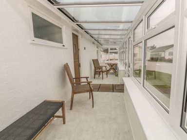A narrow enclosed porch with wooden chairs a bench a wooden door and large windows at Ty Taid in Rhosneigr